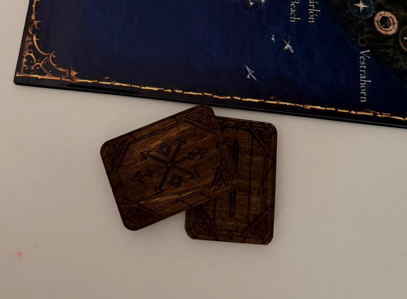 Dos fichas de juego de mesa de madera teñida, grabadas a láser con marco y símbolo, junto a la esquina del tablero de su juego